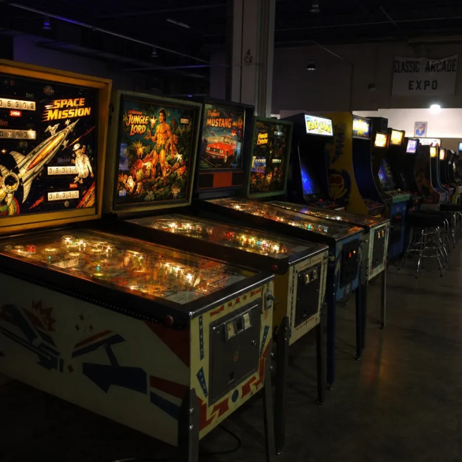 A line of vintage pinball machines and arcade cabinets glowing in a dimly lit event hall